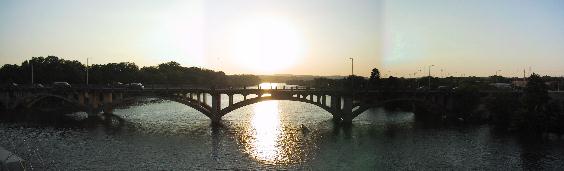 The Sun Sets behind the full span of a multi-arch bridge, the Sun Reflects on the water as a rowing team passes under the bridge.  LAMAR STREET BRIDGE from the East side.